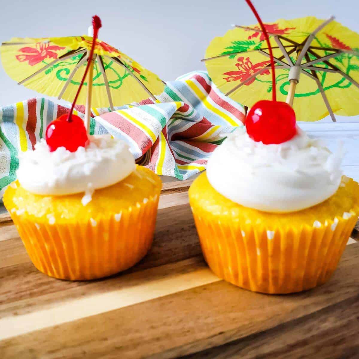 Two pina colada cupcakes with cherries on top sitting on a cutting board.