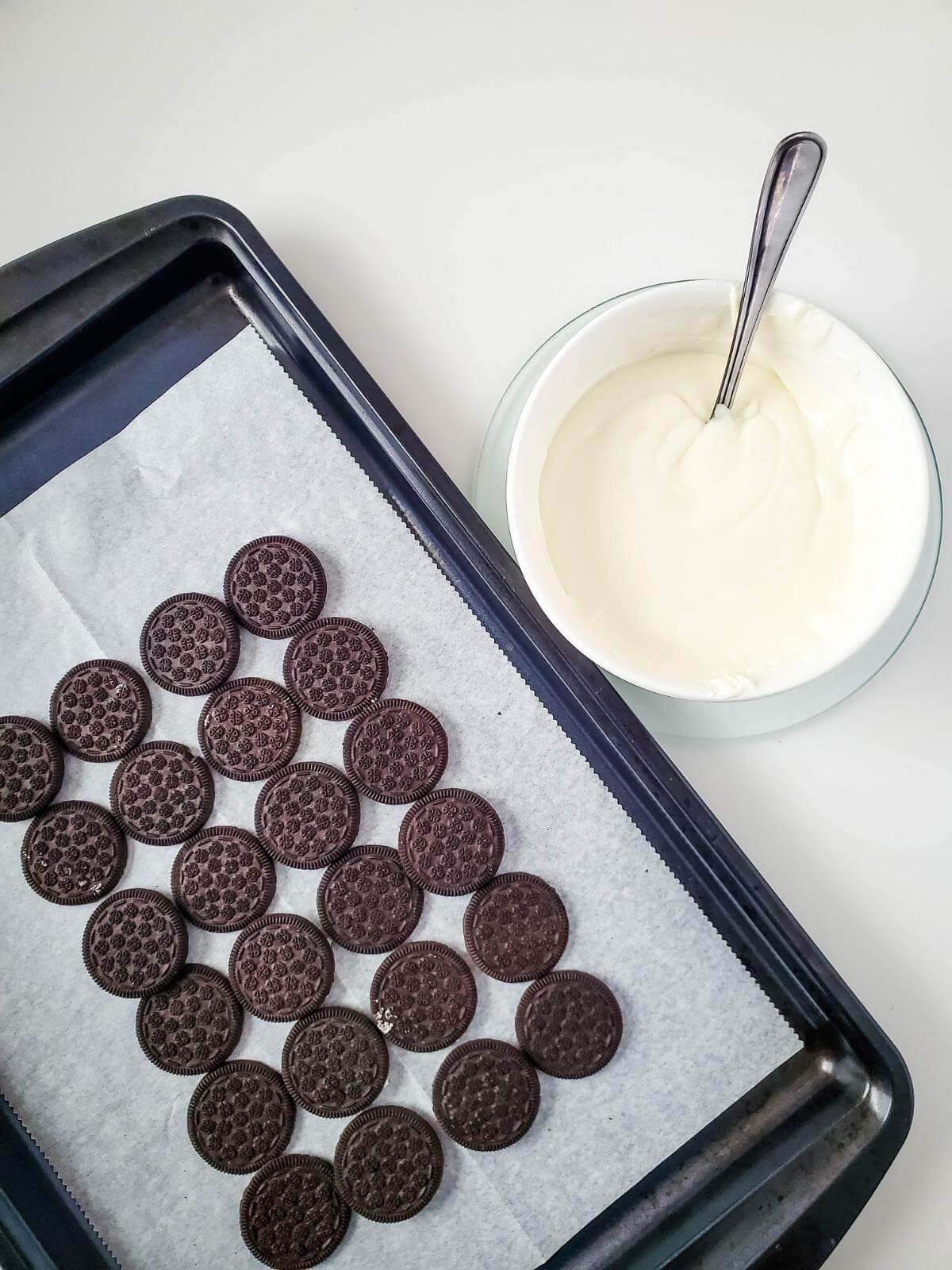 Oreos on a baking sheet next to a bowl of melted white chocolate.